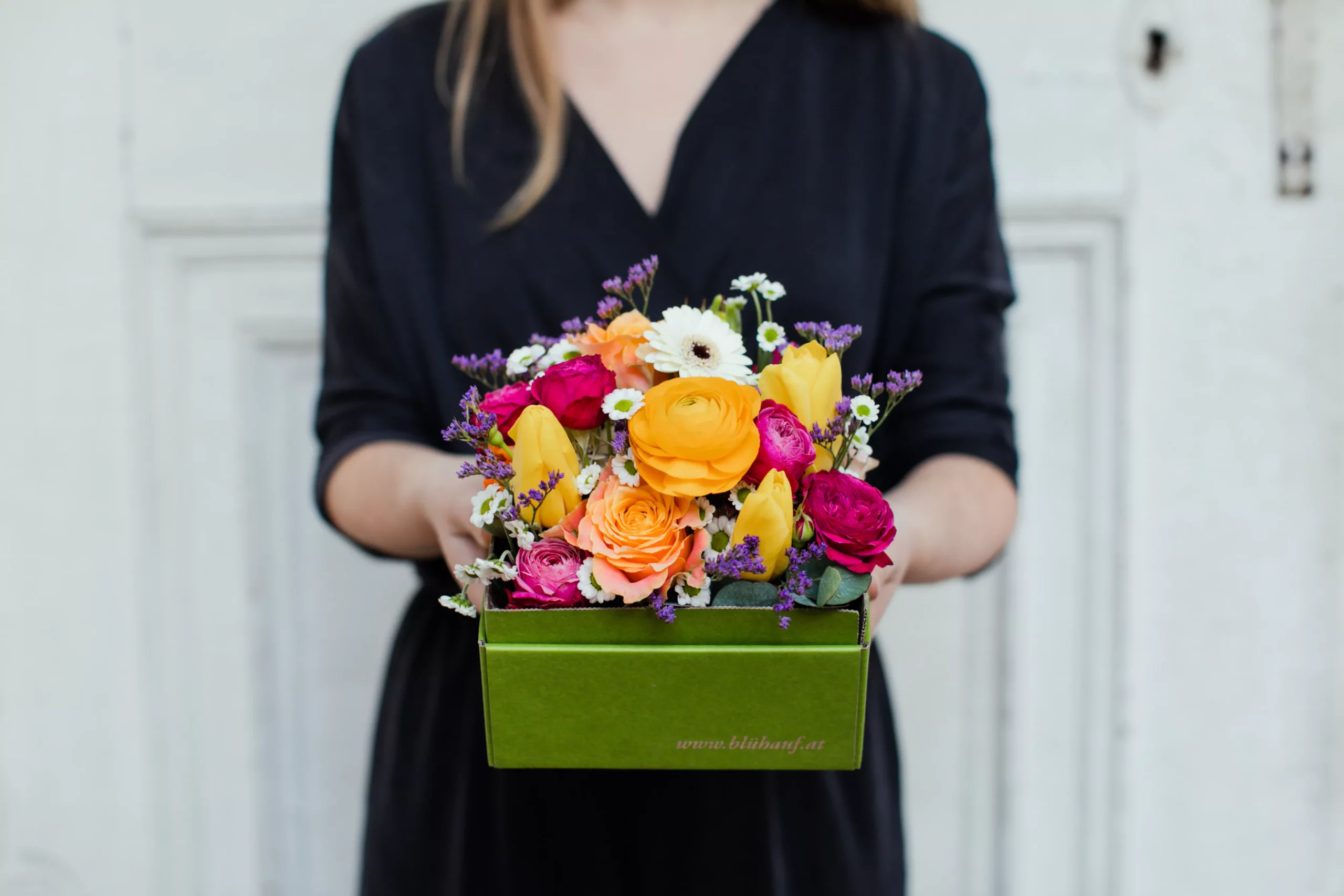 Eine Frau in einem schwarzen Kleid hält eine Blumenbox mit einer bunten Mischung aus Blumen